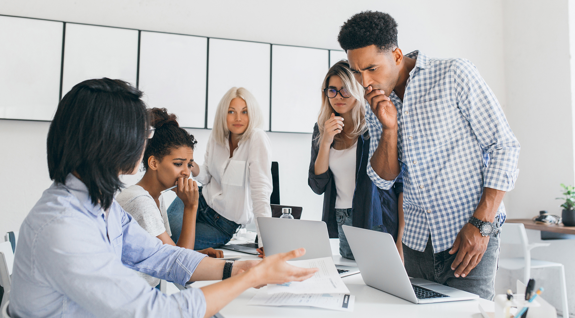 Pensive african office worker covering mouth with hand, while solving problem with computer. Team of asian and black web-programmers found a mistake in their project..