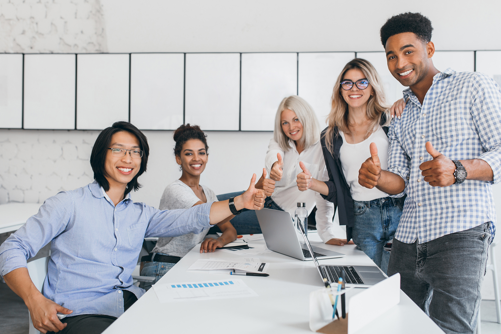 Blonde secretary sitting on table while office workers posing with thumbs up. Indoor portrait of happy asian manager in trendy shirt smiling in conference hall with foreign partners..